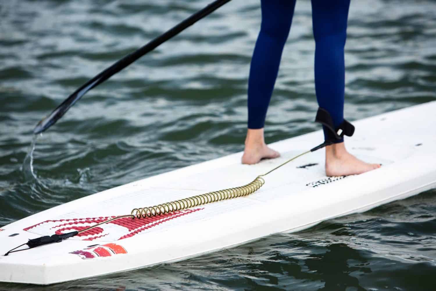 Close up picture of persons feet on a paddle board. White paddle board, black paddle, and navy blue dry suit in Clipper Cove.